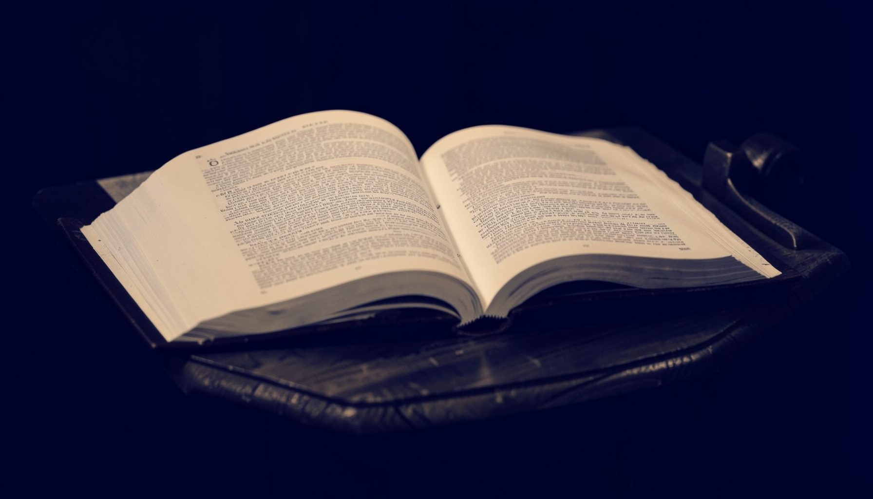 Warm duotone photograph of an open Bible resting on a wooden lectern, deep navy and amber tones